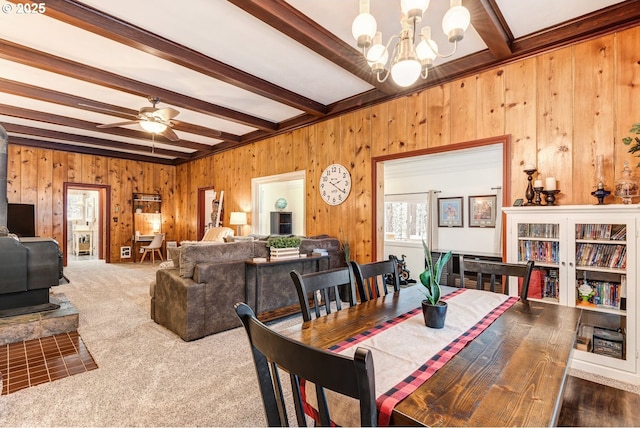 carpeted dining space with beam ceiling, ceiling fan with notable chandelier, wood walls, and a wood stove