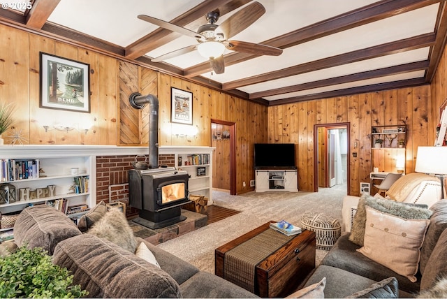 carpeted living room with a wood stove, wooden walls, a ceiling fan, and beam ceiling