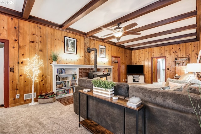 carpeted living room with beam ceiling, wooden walls, ceiling fan, and a wood stove