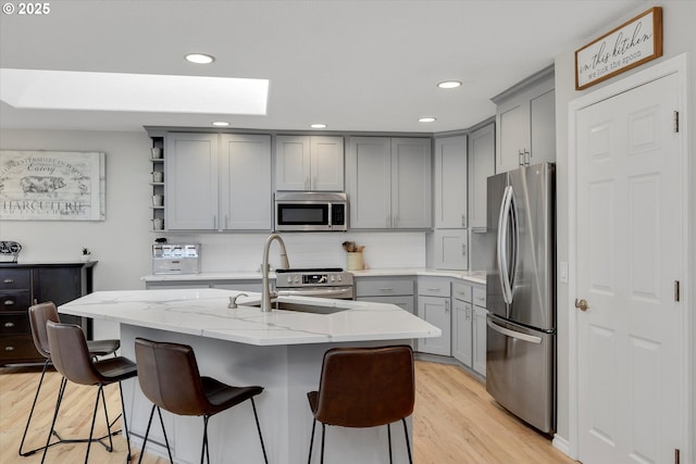 kitchen featuring light wood finished floors, a center island with sink, appliances with stainless steel finishes, light stone countertops, and open shelves