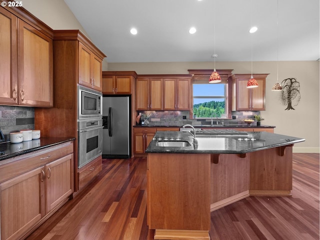 kitchen featuring dark stone counters, stainless steel appliances, sink, decorative light fixtures, and an island with sink