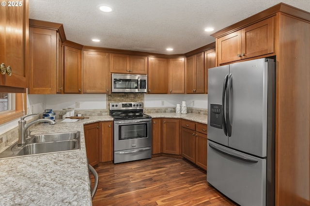 kitchen with a textured ceiling, a sink, appliances with stainless steel finishes, brown cabinetry, and dark wood finished floors