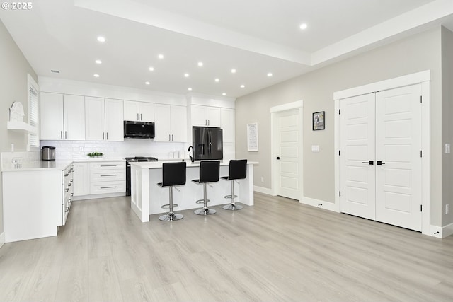 kitchen featuring a kitchen bar, a kitchen island with sink, black fridge, white cabinetry, and light wood finished floors