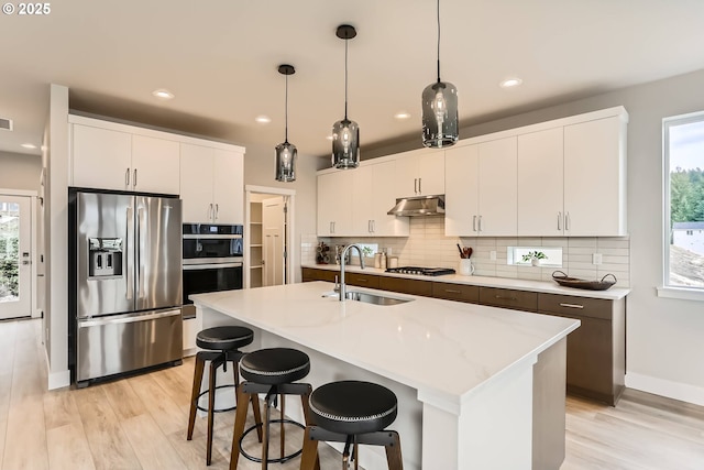 kitchen with a kitchen island with sink, stainless steel appliances, under cabinet range hood, white cabinetry, and a sink