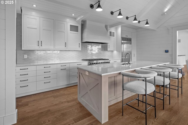 kitchen with premium range hood, white cabinetry, stainless steel built in refrigerator, stove, and a kitchen island with sink