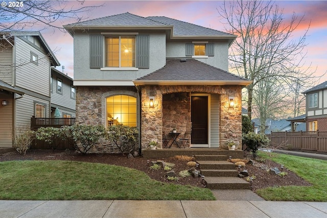 view of front of property with stucco siding, stone siding, fence, covered porch, and a shingled roof