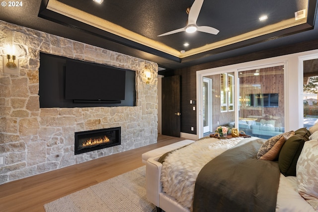 bedroom featuring light wood-type flooring, ceiling fan, a stone fireplace, and a tray ceiling