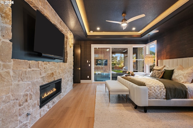 bedroom featuring hardwood / wood-style floors, a stone fireplace, ceiling fan, a tray ceiling, and access to outside