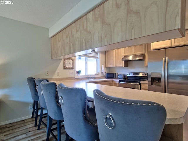 kitchen featuring a breakfast bar, stainless steel appliances, under cabinet range hood, light brown cabinets, and a sink