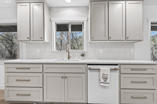 kitchen with tasteful backsplash, light stone countertops, white dishwasher, gray cabinetry, and a sink