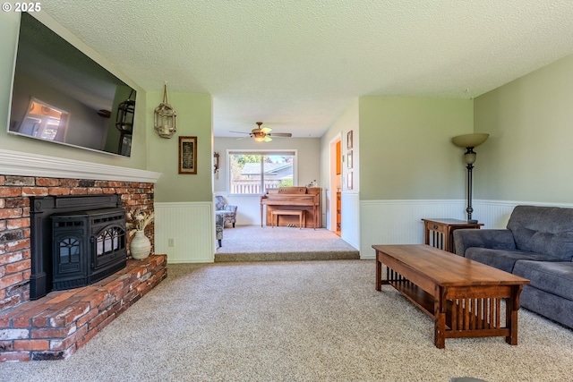 living area featuring ceiling fan, a wood stove, carpet floors, a textured ceiling, and a wainscoted wall