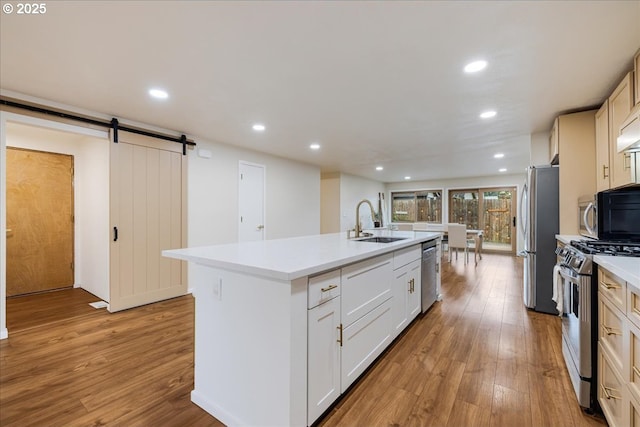 kitchen featuring a barn door, an island with sink, stainless steel appliances, light wood-type flooring, and a sink