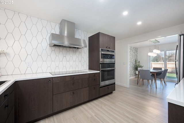 kitchen featuring pendant lighting, backsplash, wall chimney range hood, light hardwood / wood-style flooring, and stainless steel appliances