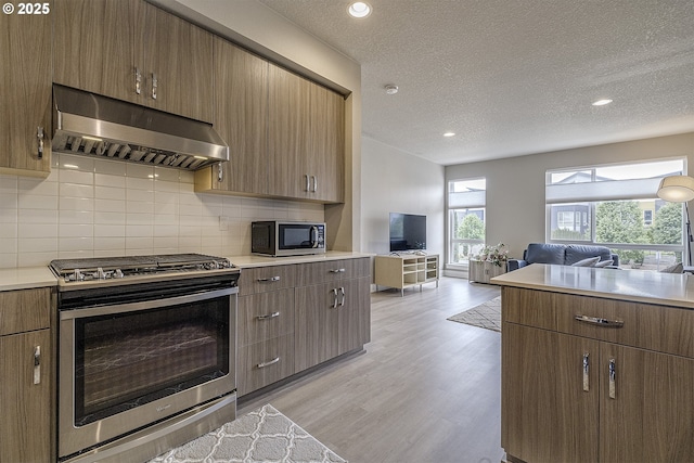 kitchen with stainless steel appliances, open floor plan, decorative backsplash, light wood-type flooring, and under cabinet range hood