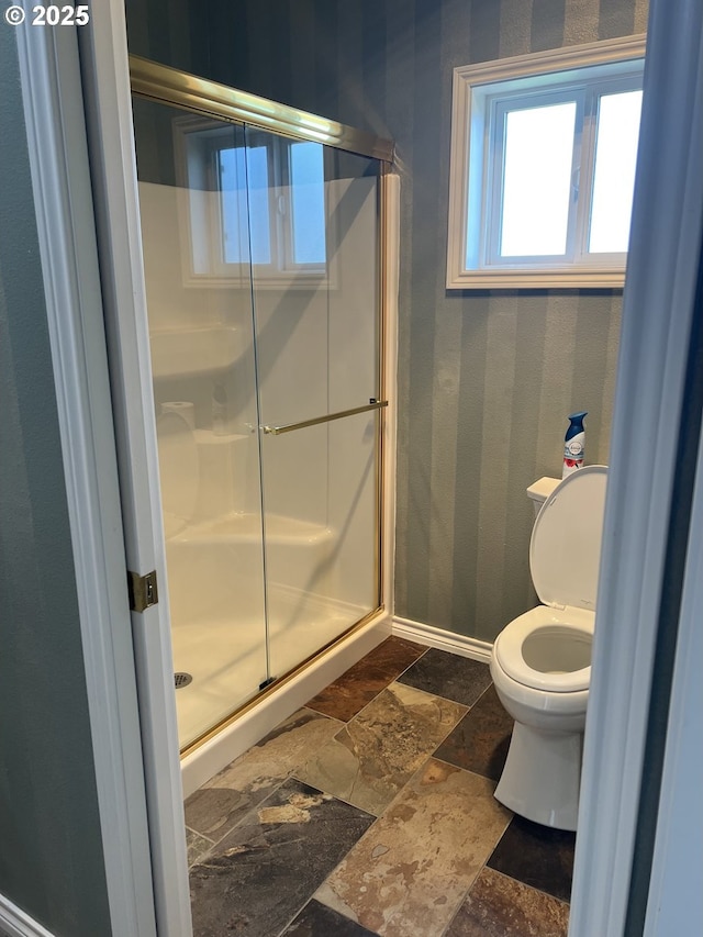 bathroom with stone tile flooring, a stall shower, toilet, baseboards, and wallpapered walls