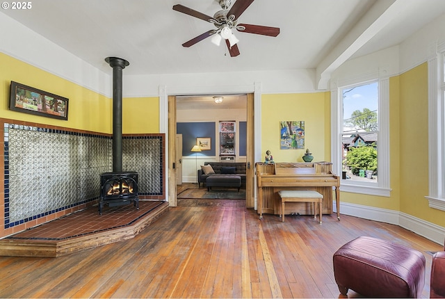 sitting room featuring a wood stove, hardwood / wood-style flooring, ceiling fan, and baseboards