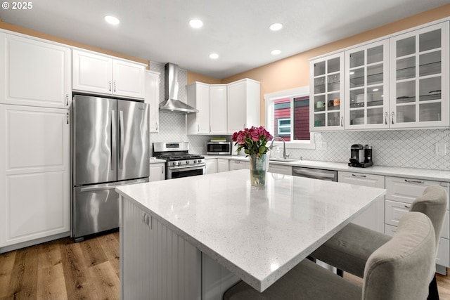 kitchen with appliances with stainless steel finishes, wall chimney range hood, white cabinets, glass insert cabinets, and light wood-style flooring