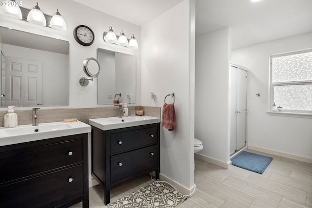 bathroom with toilet, two vanities, a shower stall, and baseboards