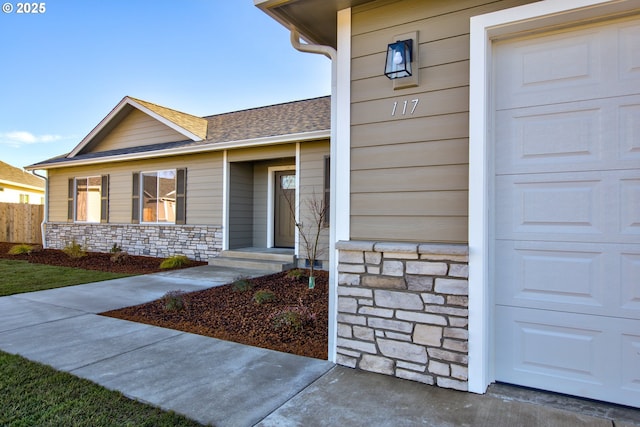 view of exterior entry with a garage, fence, stone siding, and roof with shingles