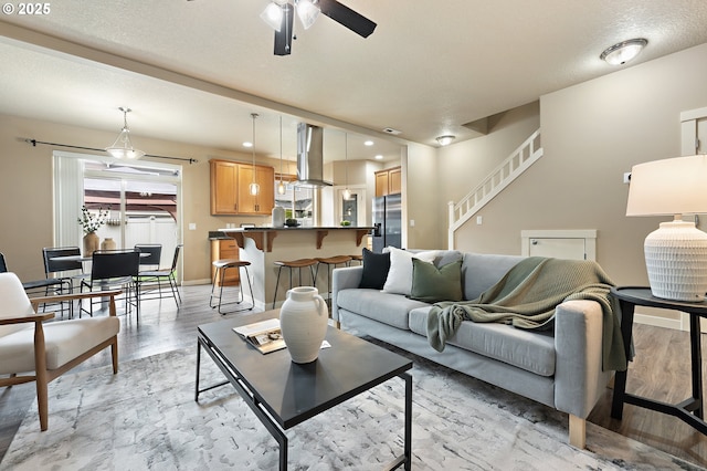 living area with baseboards, a ceiling fan, stairs, a textured ceiling, and light wood-type flooring