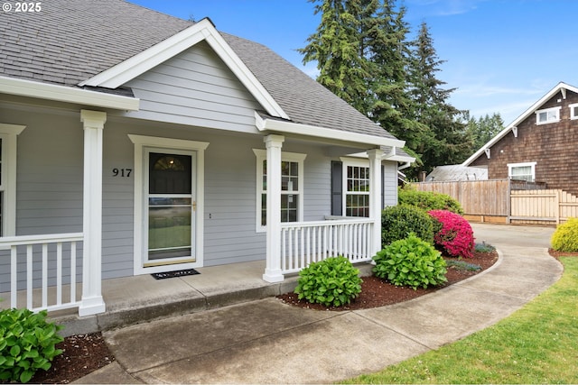 view of exterior entry featuring roof with shingles, covered porch, and fence