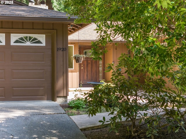 doorway to property with an attached garage, roof with shingles, and driveway