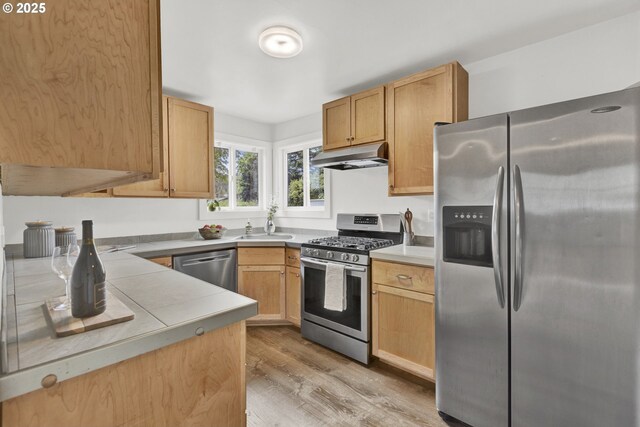 kitchen with stainless steel appliances, under cabinet range hood, light wood-style flooring, a sink, and light brown cabinetry