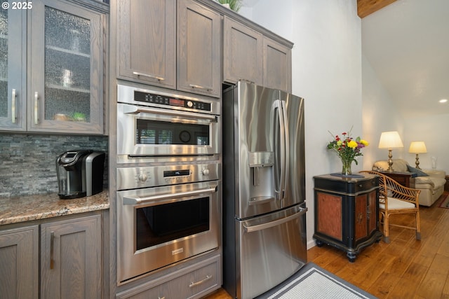 kitchen featuring decorative backsplash, glass insert cabinets, wood finished floors, light stone countertops, and stainless steel appliances