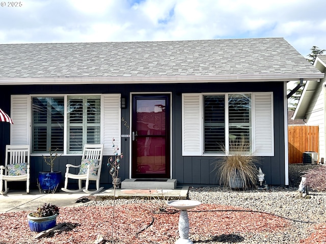 view of exterior entry with roof with shingles, covered porch, central AC unit, board and batten siding, and fence