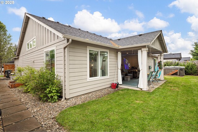 back of house featuring roof with shingles, a hot tub, board and batten siding, a lawn, and a patio