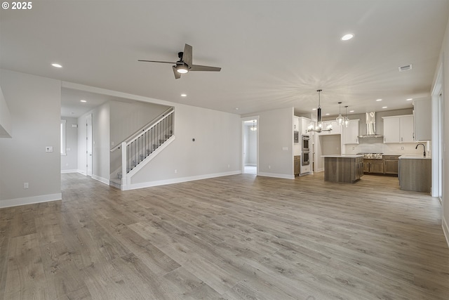 unfurnished living room featuring stairs, light wood-type flooring, recessed lighting, ceiling fan with notable chandelier, and visible vents