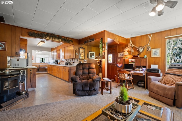 living room featuring wooden walls, a desk, a ceiling fan, a wood stove, and light tile patterned flooring