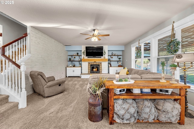 carpeted living room with a warm lit fireplace, ceiling fan, a textured ceiling, and stairs