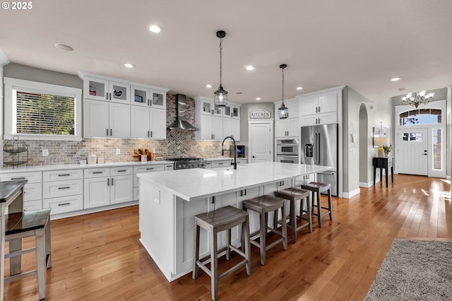 kitchen featuring arched walkways, stainless steel appliances, light wood-style flooring, a sink, and wall chimney exhaust hood