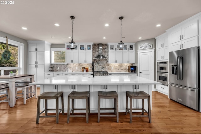 kitchen featuring white cabinetry, light countertops, appliances with stainless steel finishes, wall chimney exhaust hood, and glass insert cabinets