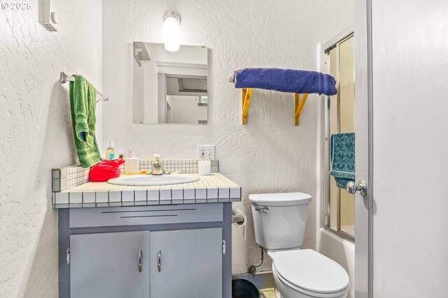 full bathroom with combined bath / shower with glass door, toilet, vanity, and a textured wall