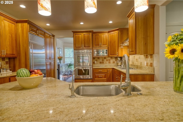 kitchen with stainless steel appliances, brown cabinetry, a sink, and light stone countertops