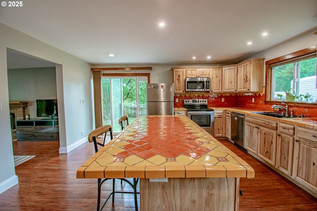 kitchen featuring stainless steel appliances, tile countertops, light brown cabinetry, a sink, and decorative backsplash