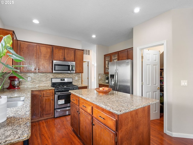 kitchen with appliances with stainless steel finishes, light stone counters, decorative backsplash, dark wood-type flooring, and a kitchen island