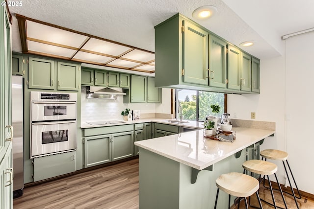 9080 NW Sherry Ct, Portland, OR 97229 - kitchen with green cabinetry, stainless steel appliances, a peninsula, under cabinet range hood, and light wood-type flooring