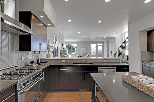 kitchen with appliances with stainless steel finishes, wall chimney range hood, a sink, decorative backsplash, and recessed lighting
