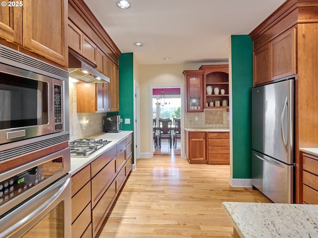 kitchen featuring backsplash, stainless steel appliances, exhaust hood, an inviting chandelier, and light hardwood / wood-style floors