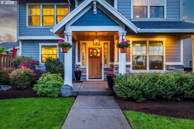 exterior entry at dusk featuring roof with shingles, fence, covered porch, and stone siding
