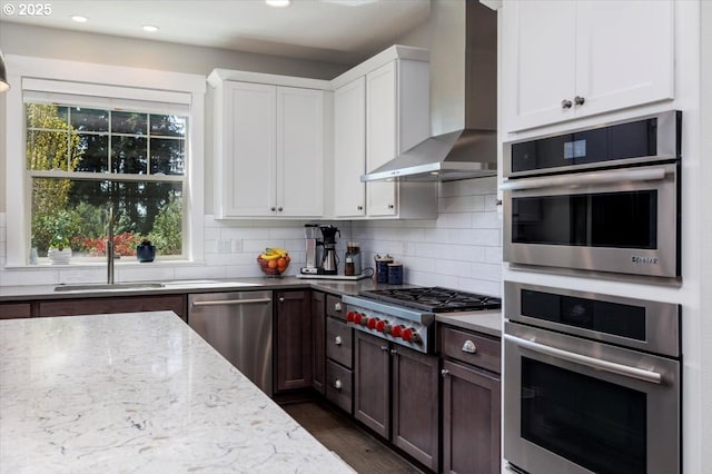 kitchen with white cabinets, light stone counters, wall chimney exhaust hood, stainless steel appliances, and tasteful backsplash