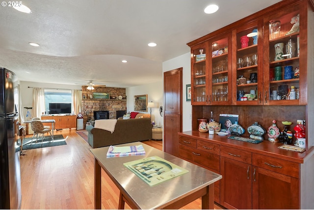 kitchen featuring stainless steel countertops, light wood-style flooring, freestanding refrigerator, ceiling fan, and a stone fireplace