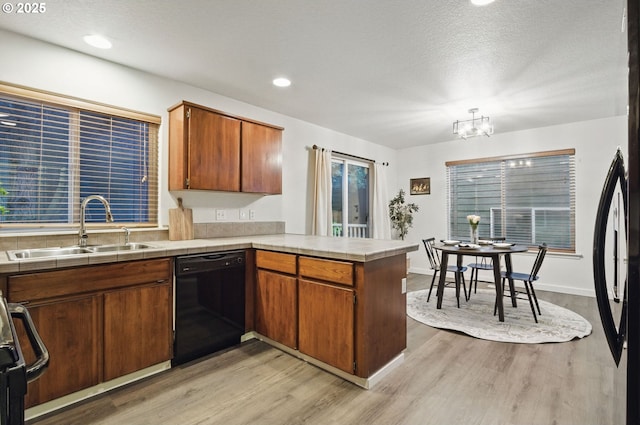 kitchen featuring a peninsula, a sink, light wood-type flooring, black dishwasher, and tile counters