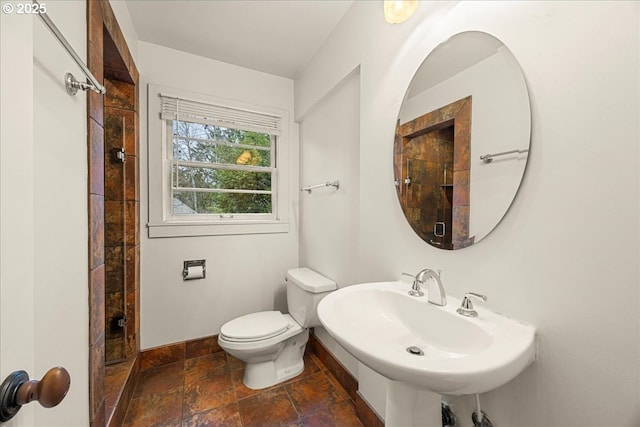 bathroom featuring toilet, stone tile floors, a sink, baseboards, and a tile shower