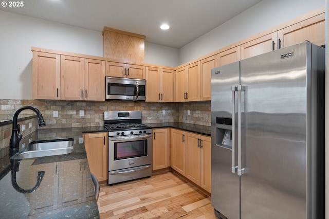 kitchen featuring appliances with stainless steel finishes, a sink, light brown cabinetry, light wood-style floors, and dark stone countertops