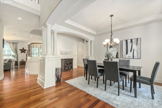 dining area with a notable chandelier, wood finished floors, arched walkways, decorative columns, and a raised ceiling