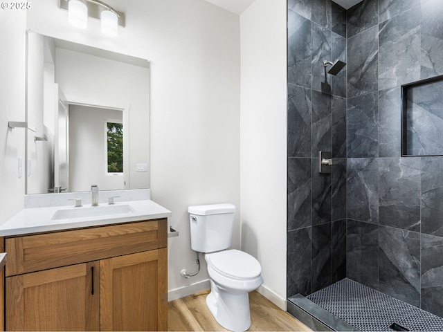 bathroom featuring toilet, wood finished floors, tiled shower, baseboards, and vanity